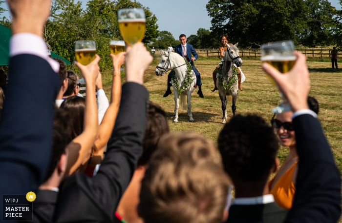 In Hertfordshire, UK, the newlyweds make a grand entrance at the reception on horseback, greeted warmly by guests who joyfully raise their glasses in celebration and welcome.