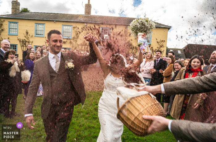 In Hertfordshire, UK, during the lively confetti toss, a full basket's worth of colorful confetti is joyfully aimed toward the bride's face, creating a playful and cheerful scene on the lawn.