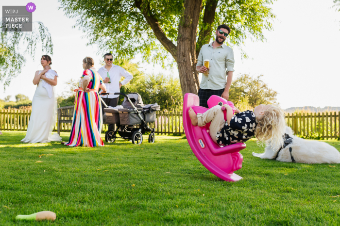 Young Guest Tests Rocking Horse's Limits, Captivating Men, at Sopley Mill Gardens In the vibrant gardens of Sopley Mill, a young guest eagerly tests the limits of a rocking horse, captivating the attention of the men, as the women chat animatedly amongst themselves.