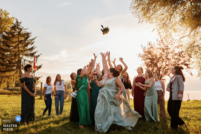 Eager Guests Gather for Bouquet Toss under Sunlight at Cocktail Reception in Fribourg, Switzerland In Fribourg, Switzerland, under the wonderful sunlight, an eager group gathers on the grass for the exciting bouquet toss, adding lively anticipation to the cocktail reception.