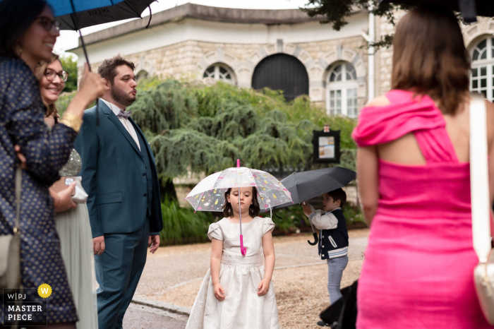 Young Girl Balances Umbrella on Her Head in Whimsical Scene During Rainy Ceremony in Oise, Hauts-de-France In the quaint setting of Oise, Hauts-de-France, a young girl cleverly supports an umbrella with her head, creating a charming and whimsical scene amidst the gentle rain during the ceremony.