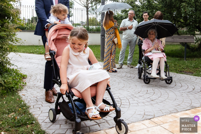 Elderly Woman in Wheelchair Watches Children Play with Stroller at Île-de-France Ceremony At a ceremony in Île-de-France, an elderly woman in a wheelchair watches warmly as children play nearby with a stroller, filling the air with joyful laughter and energy.