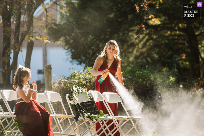 At a private residence in Strasburg, VA, a junior bridesmaid diligently sprays mosquito repellent around the ceremony area, ensuring the flower girl remains undisturbed by the buzzing pests.