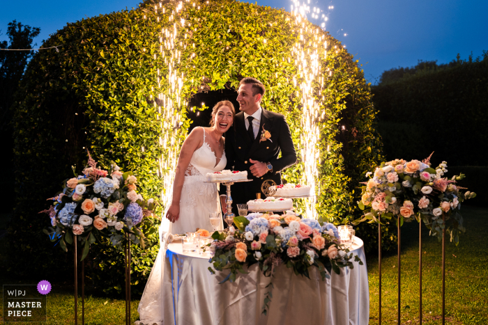 Bride and Groom Prepare to Cut Cake Under Vibrant Fireworks at Castello Bevilacqua, Verona, Italy At Castello Bevilacqua, Verona, Italy, the smiling bride and groom prepare to cut their cake, their joy illuminated by vibrant fireworks bursting in the dusk sky behind them.