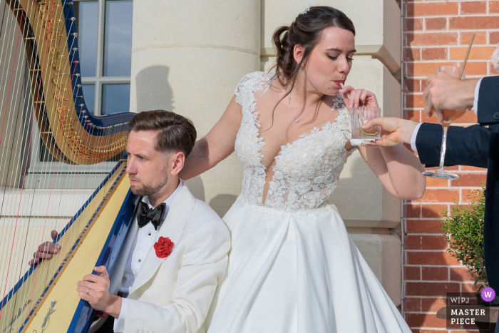 At the Versailles Yvelines reception venue, the bride and groom enjoy a unique moment during the cocktail hour. The bride sips her drink through a straw, while the groom sits, holding a harp, creating a charming scene.