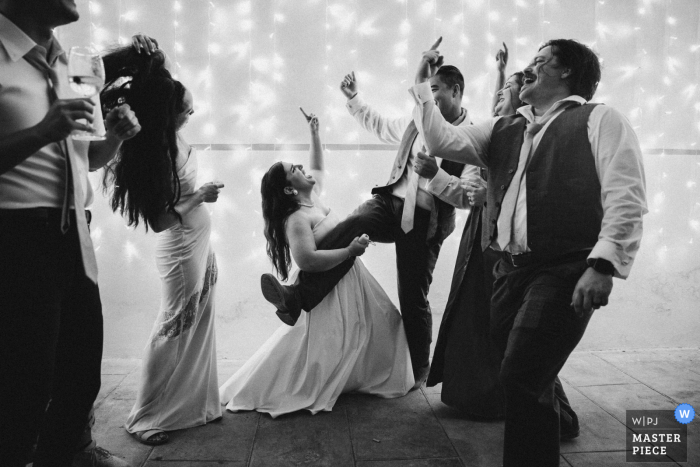Bride Rocks Air Guitar on Guest's Leg in Black-and-White Photo at Villa Orlando Dance Party At Villa Orlando in Italy, during a lively rock 'n roll moment captured in black and white, the bride playfully strums an air guitar on a man's leg. This energetic and fun-filled scene encapsulates the spirited atmosphere of the dance party.