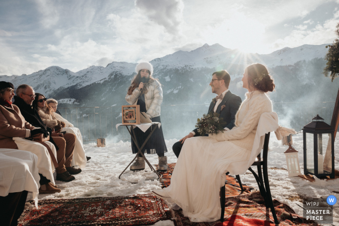 Bride and Groom Face Guests at Secular Ceremony in Valais, Sunlit Swiss Alps Creating Snowy Scene In Valais, Switzerland, a secular ceremony unfolds in the Swiss Alps with the bride and groom facing their seated guests. The low sun peeks over the mountains, illuminating the brisk, snowy scene.