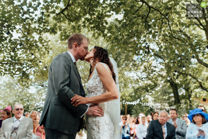 At Hothorpe Hall in Leicestershire, UK, the bride and groom share their first kiss beneath a full canopy of trees, surrounded by the beauty of the woods and their guests.