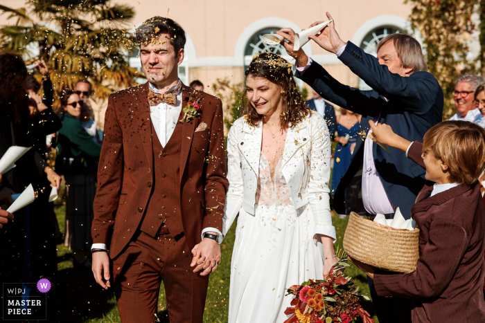 Bride and Groom Depart Ceremony in Fribourg Under Sunlit Petal Shower In Fribourg, Switzerland, the bride and groom depart the ceremony amidst a shower of dried petals, illuminated by natural sunlight.