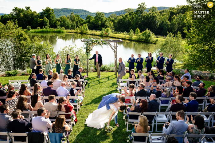 Couple's Joyful Recessional Kiss at The Ponds at Bolton Valley At The Ponds at Bolton Valley in Bolton, VT, a high-angle shot captures the couple kissing and recessing from the ceremony on the grass. Sunlight bathes the scene, highlighting the joy and natural beauty of their special day.