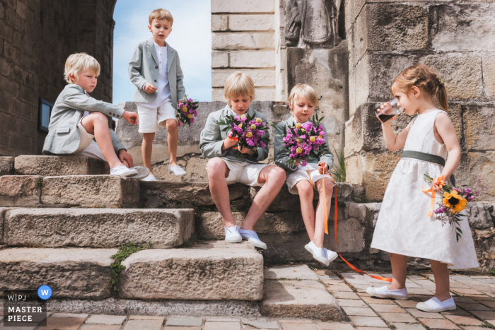 At Eglise d'Urrugne in Pyrénées Atlantiques, France, the photographer captures the young attendants waiting patiently on the church steps after the religious ceremony, highlighting composed innocence and skillful storytelling.