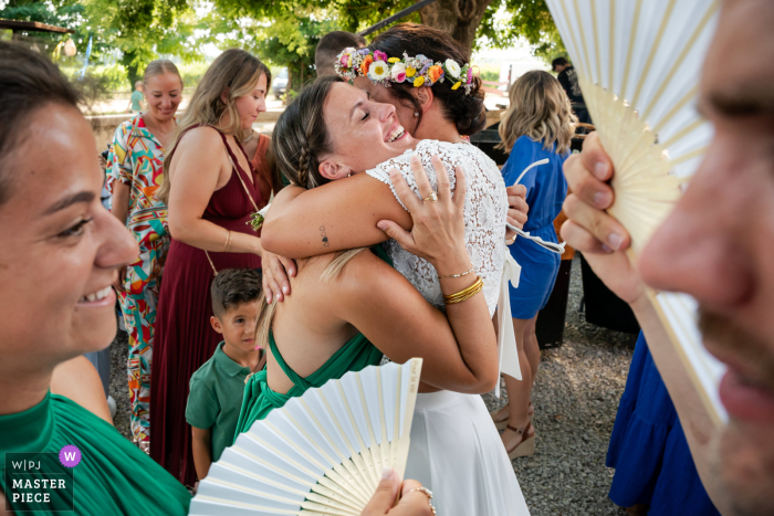 In Mauguio, France, the photographer artistically captures the bride hugging a guest, beautifully framed by others in the foreground, showcasing skillful composition and an authentic celebration of friendship.