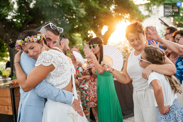 Golden Hour Embrace: A Celebration of Love and Luminous Light In Mauguio, France, the photographer captures the bride and groom hugging beneath sunlit trees, with another embracing duo at right, masterfully highlighting warmth, layered emotion, and luminous natural light.
