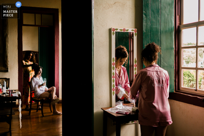 In São Caetano do Sul, São Paulo, one woman sits in a distant room having her hair styled, while another does her makeup in the mirror in the foreground, illustrating the layered preparations before the wedding.