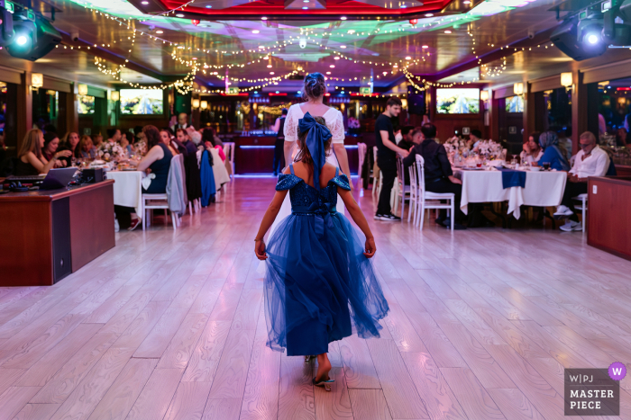A whimsical moment in Istanbul, Turkey, as a young girl in a blue dress playfully mimics the bride, walking behind her across an empty dance floor. Captured from behind, the image radiates innocence and joy.