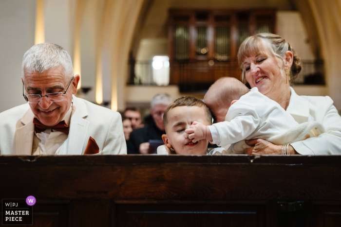 A Baby Pinches A Boy’s Nose In The Church Ceremony In Metz, France, Delighting And Amusing Wedding Guests Nearby. Inside the church, a spontaneous, lighthearted moment unfolds as a baby pinches a young boy’s nose during the ceremony, while an amused woman and sharply dressed man look on.