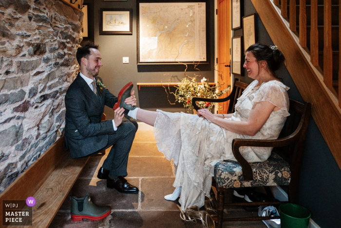   At Hause Hall Farm in Cumbria, England, the groom helps his new wife remove her boots on their wedding day, illustrating the couple’s comfort and caring in a rustic setting.