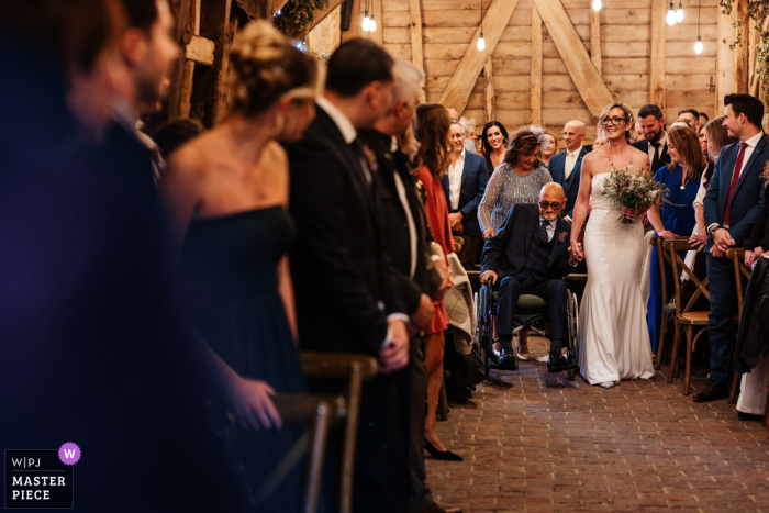 The Bride Is Walked Down The Aisle By Her Dad In A Wheelchair At High Billinghurst Farm, Surrey, England. The bride is lovingly walked down the aisle by her dad, who uses a wheelchair, at High Billinghurst Farm in Surrey, England, highlighting an emotional and supportive wedding entrance.