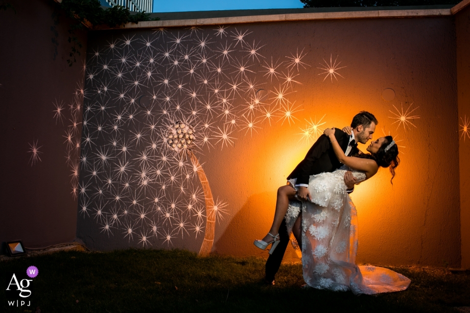 Foto de: <a href="https://www.wpja.com/photos/artistic-wedding-photographers/2558974"> Orçun Yalçın - Ankara Ankara Fotografía de la boda de Wyndham Ankara: el novio sostiene a la novia desde su cadera justo al lado de la pared con un mural de flores y resaltado con un color naranja