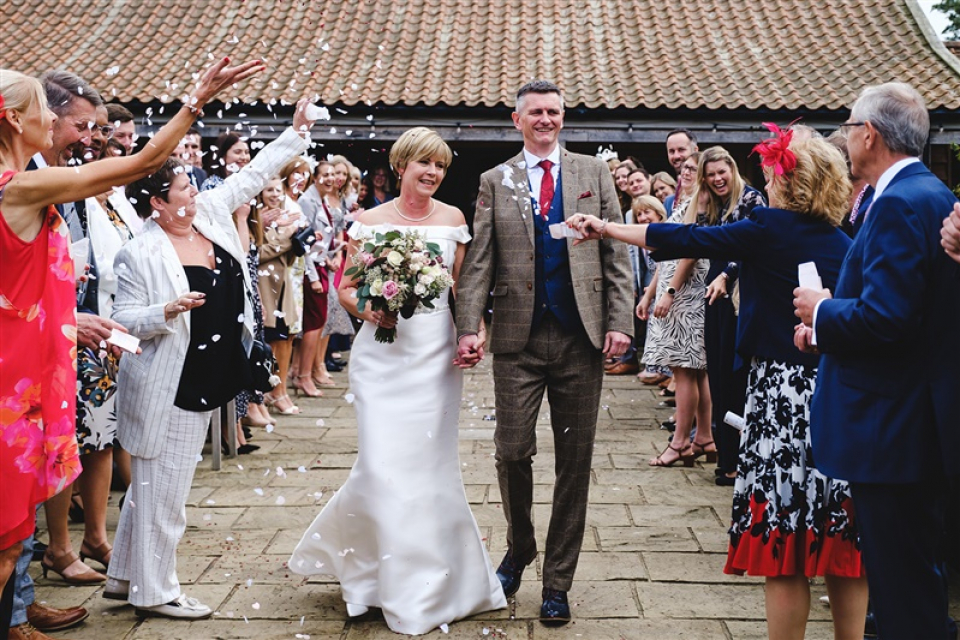 La mariée et le marié reçoivent une douche de confettis de la part de la famille et des invités sur la terrasse extérieure du lieu de mariage Easton Grange dans le Suffolk Couple couvert de confettis sur la terrasse du lieu de mariage Suffolk Easton Grange