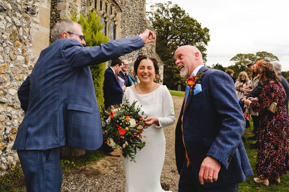 Le couple rit devant l'église de Polstead dans l'Essex sous les douches de confettis Les mariés rient et canard alors qu'ils sont couverts de confettis par un invité de mariage à l'extérieur d'une église de campagne anglaise dans le Suffolk