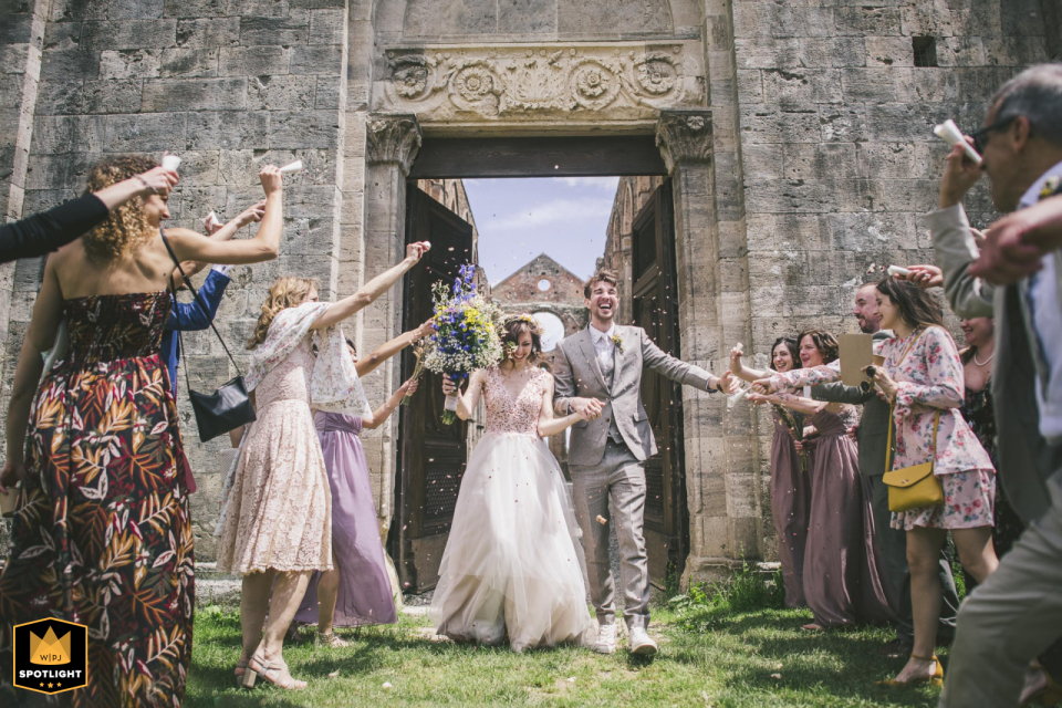 A bride and groom walk hand in hand through San Galgano Abbey ruins in Tuscany. The wedding ceremony has concluded, and the couple receive congratulations as they exit.