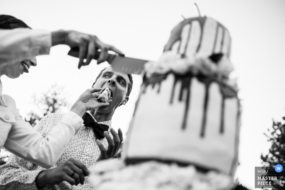Photo by: <a href="https://www.wpja.com/contests/2018-v07-photography-competition/best-wedding-photographers/2228076">Carl Bower - Denver Colorado</a> Bride wields knife and cake at outdoor wedding in Boulder, Colorado. Bride forces wedding cake into groom's mouth. Sunrise Amphitheater, Boulder, Colorado