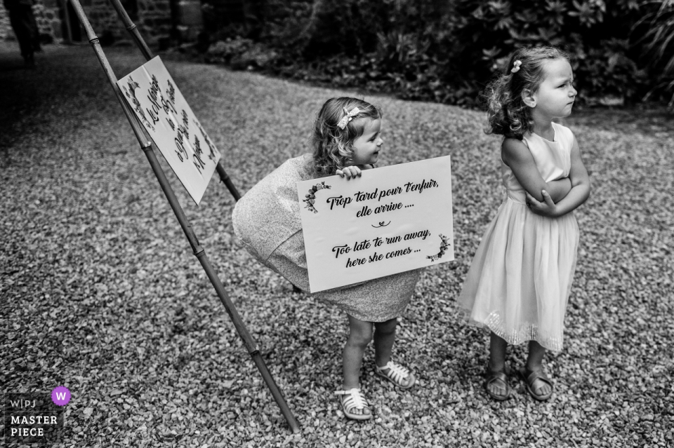 Dos niñas permanecen afuera mientras una sostiene un cartel en esta foto en blanco y negro tomada fuera de Le Clos de Trevannec en Francia por un fotógrafo de bodas en Victoria, Australia.