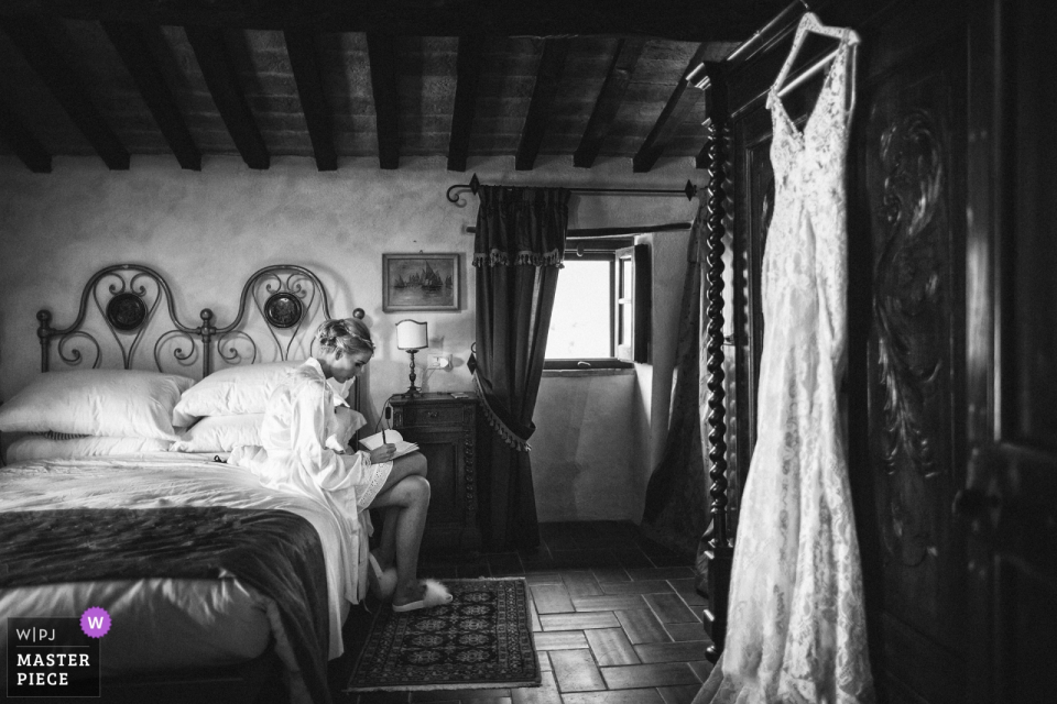 Foto di: <a href="https://www.wpja.com/contests/2018-v10-photography-competition/best-wedding-photographers/2250636"> Andrea Cittadini - Spoleto Perugia </a> Foto di nozze di Abbazia San Faustino della sposa che scrive una nota sul letto mentre il suo vestito appende nelle vicinanze.