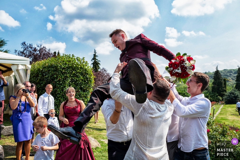 Turin Wedding Photo of the groom lifted above the men as he holds a bouquet of flowers at the Tenuta castello Cerrione Turin Wedding Photojournalist | the groom is lifted above the men as he holds a bouquet of flowers at the Tenuta castello Cerrione