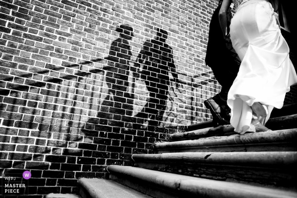 Photo by: <a href="https://www.wpja.com/contests/2018-v13/best-wedding-photographers/2290296">Johnny Shryock - Arlington Virginia</a> Baltimore Country Club wedding photograph of the bride and groom climbing steps with their shadows on a brick wall.