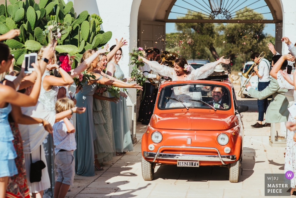 Foto por: <a href="https://www.wpja.com/contests/2018-v17/best-wedding-photographers/2340241"> Mark Wallis - Belper Derbyshire </a> Una pareja conduce a través de una línea de invitados de bodas lanzando confeti en un automóvil Fiat vintage en Masseria Potenti en Italia
