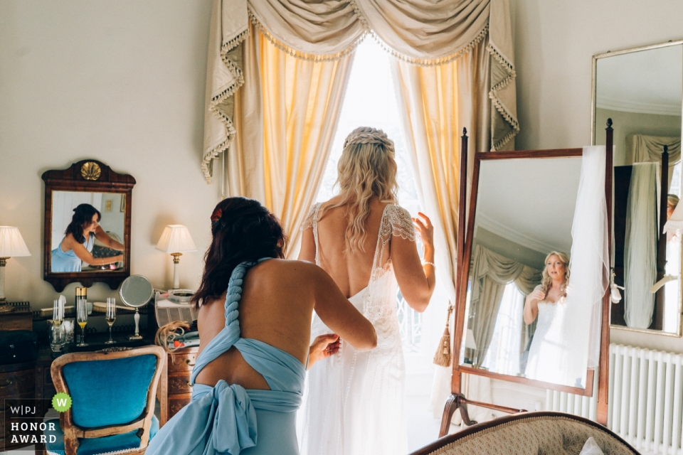 Wedding getting ready photo shoot in North Cadbury Court, Somerset UK of Bridesmaid helping the bride into wedding dress with reflections in mirrors Wedding getting ready photo shoot in North Cadbury Court, Somerset UK - Bridesmaid helping bride into wedding dress with reflections in mirrors
