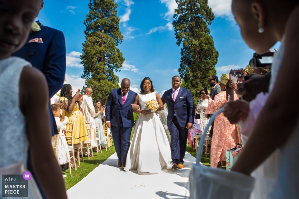 The bride is escorted down the aisle for her outdoor wedding at the Kingston Bagpuize House, London 