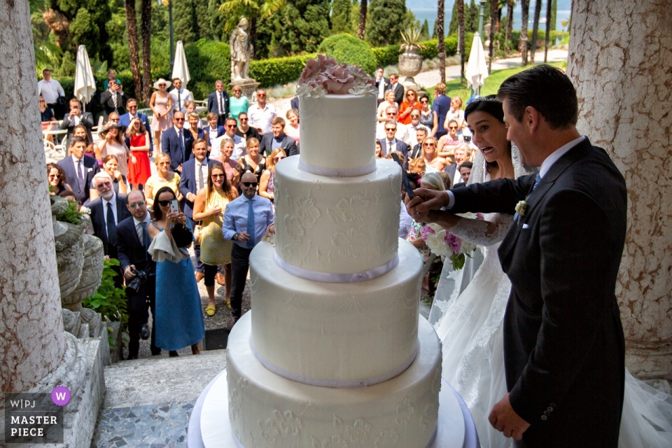 Huwelijksfotografie van buiten in Villa Cortine Palace Hotel - Sirmione - Gardameer - Italië - Afbeelding van grappig taartsnijden met de gasten op de achtergrond