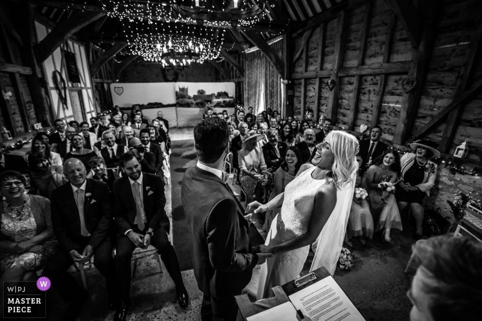 Manor Farm Barn Fotografia del luogo del matrimonio, Bicester, Oxfordshire - Sposa e sposo durante la cerimonia, la sposa improvvisamente ride ad alta voce durante l'intera cerimonia