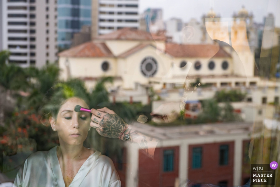 Photo de mariage du Belmond Hotel Miraflores, Lima, Pérou | Reflet dans la fenêtre de la mariée se maquillant, derrière la fenêtre est l'église dans laquelle elle est sur le point de se marier