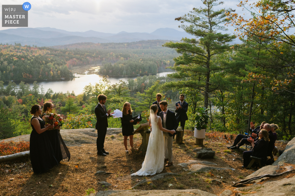 Forest Lake Camp in Chestertown, NY, afbeelding van een stel dat elkaar omhelst aan het einde van hun ceremonie op de kant van een berg terwijl de zon laag aan de hemel komt. Hun huwelijksfeest en ouders omringen hen.