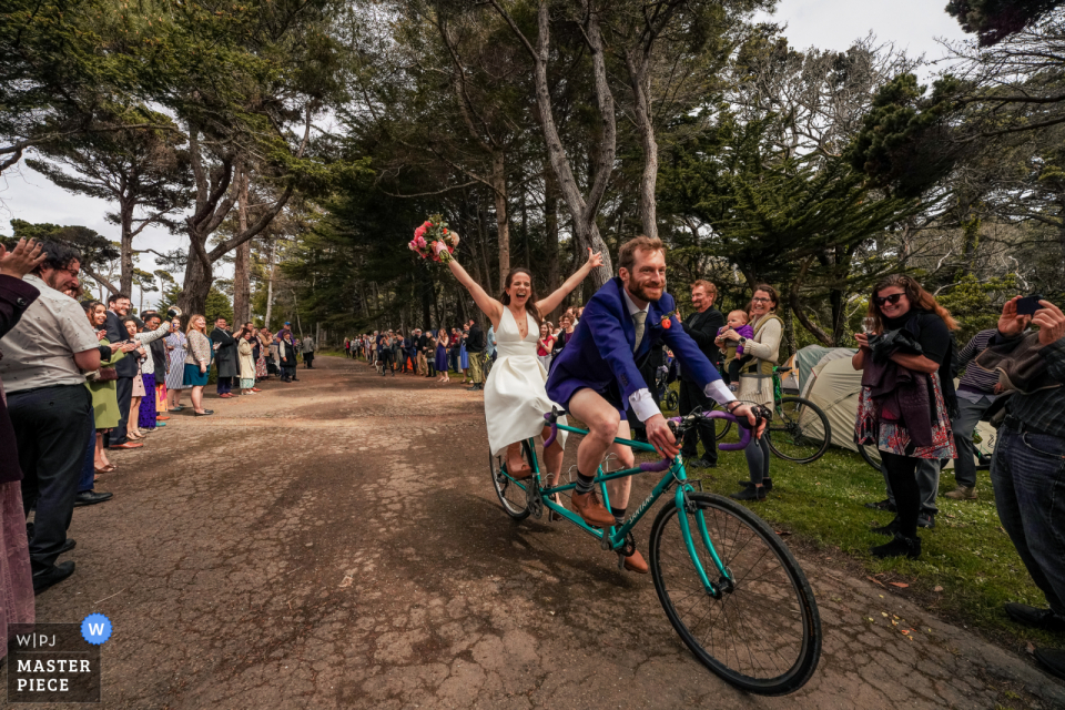 Der dokumentarische Hochzeitsfotograf am Veranstaltungsort Holly's Ocean Meadow in Fort Bragg, Kalifornien, hat dies festgehalten. Die Braut und der Bräutigam reiten auf einem Tandem vor ihren jubelnden Gästen zu ihrem Empfang Der dokumentarische Hochzeitsfotograf am Veranstaltungsort Holly's Ocean Meadow in Fort Bragg, Kalifornien, hat dies festgehalten. Die Braut und der Bräutigam reiten auf einem Tandem vor ihren jubelnden Gästen zu ihrem Empfang