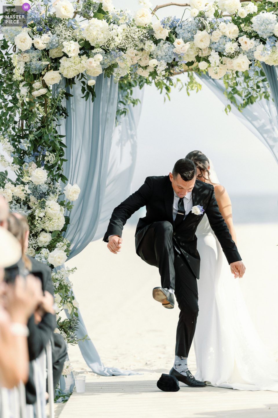 En esta foto tomada en Wychmere Beach Club en Harwich, MA, durante una ceremonia de boda judía, el novio es capturado en un momento crucial cuando está a punto de romper un vaso envuelto con su talón, simbolizando el final de la ceremonia.