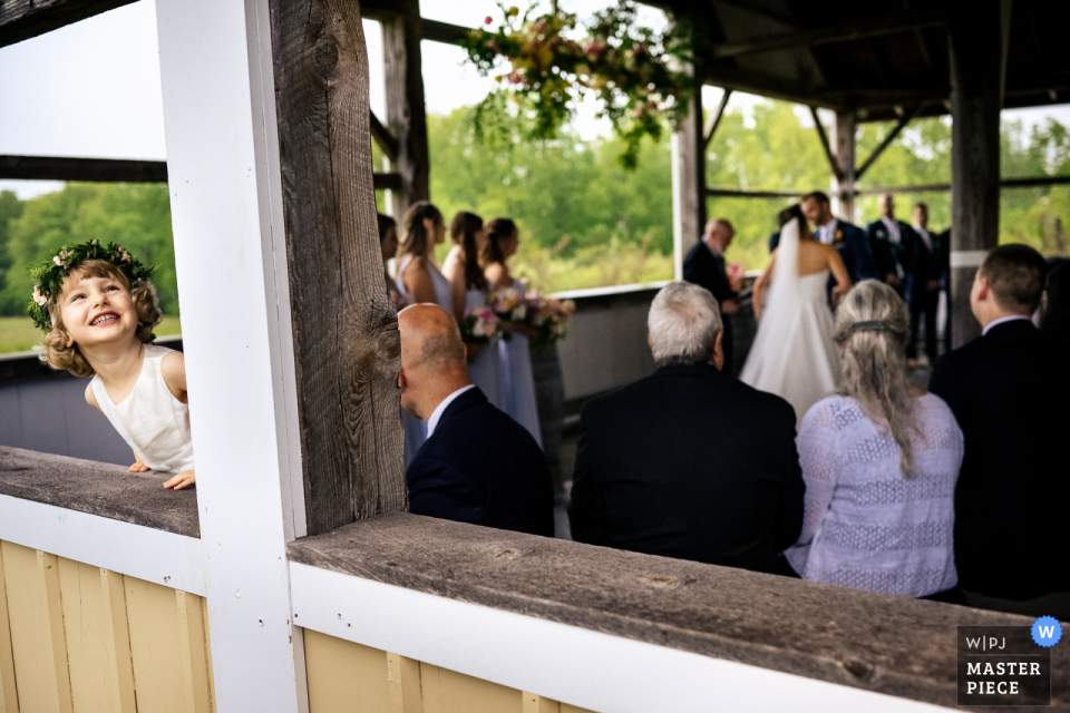 Hochzeitsfoto der Valley View Farm: Ein Blumenmädchen blickt durch offene Fenster eines verandaähnlichen Raums in Haydenville, MA, und prüft, ob es während der Zeremonie noch regnet