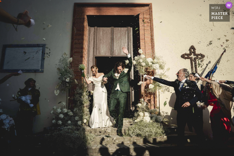 At Castello di Badia Berardenga in Tuscany, the newlyweds exit the church happily. They're showered with tossed petals in the bright sunshine after the ceremony.