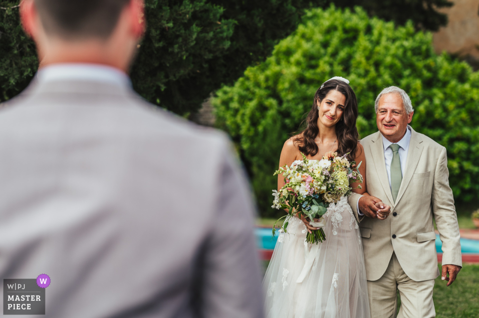 At Villa di Ulignano in Tuscany, Italy, the bride and her father share a reaction of joy and excitement upon seeing the groom as they arrive at the wedding ceremony. This heartfelt moment underscores the anticipation and happiness of the day.