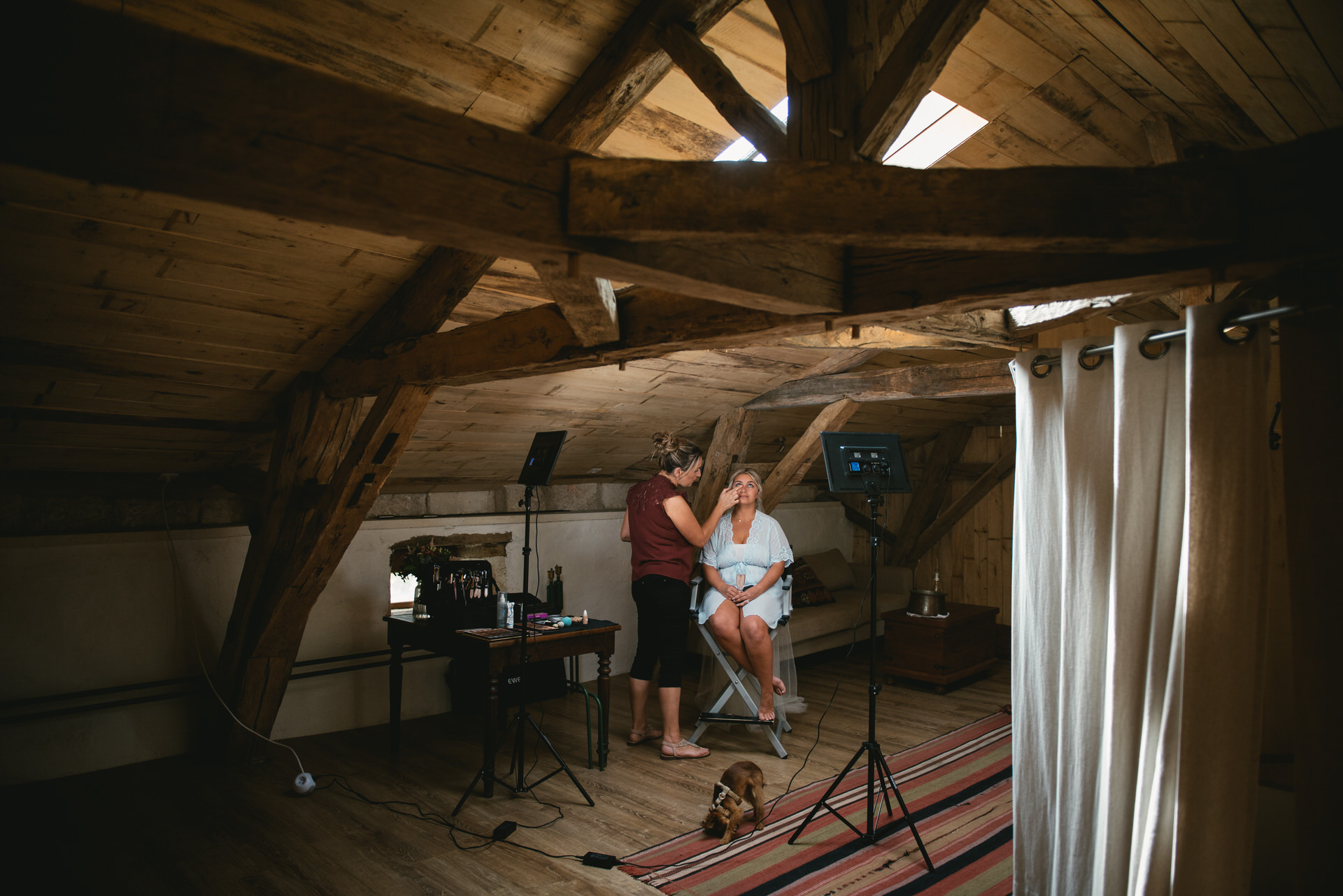 In the softly lit attic of a building in Dordogne, France, a makeup artist diligently applies makeup on a bride. The intimate setting, with rustic wooden beams and gentle light filtering in, creates a serene ambiance. 