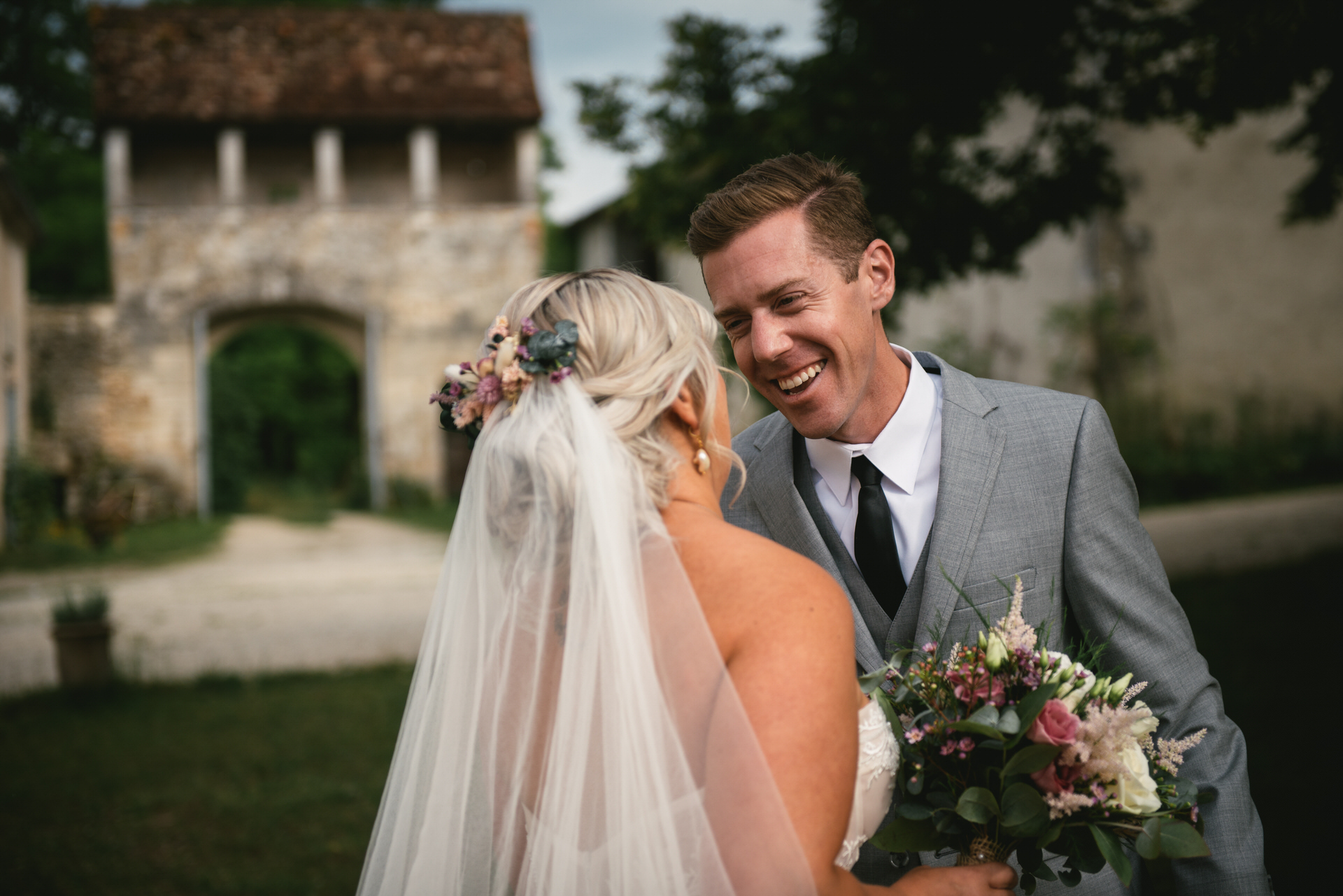 In Dordogne, France, the groom joyfully laughs with the bride during their first look. She holds a vibrant bouquet, and her veil is adorned with delicate flowers, adding a touch of elegance to the intimate moment.