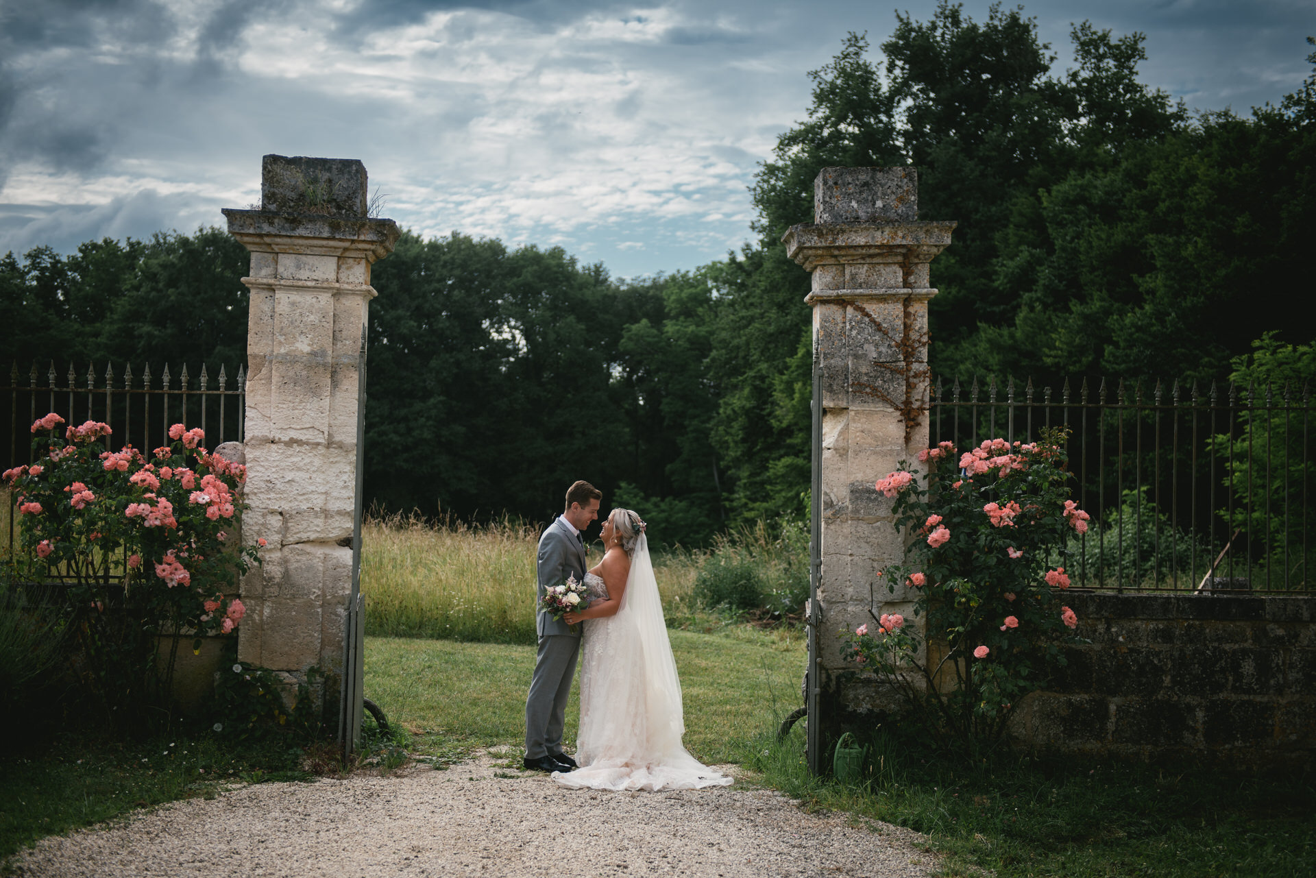 A couple shares a tender kiss beneath a stone archway adorned with blooming pink roses in Dordogne, France. They are surrounded by the serene beauty of nature, creating an intimate and romantic atmosphere for their elopement.