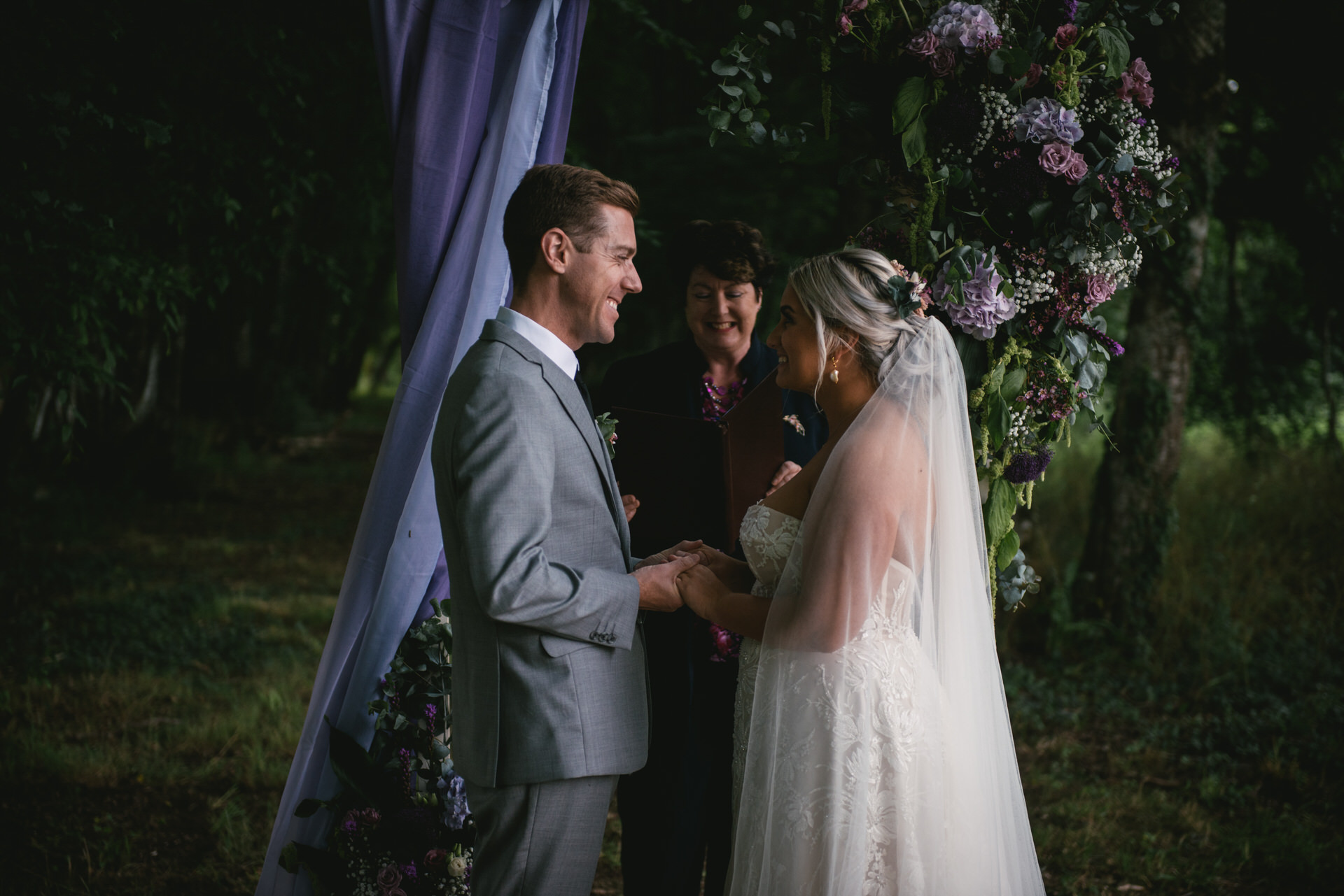 A joyful couple holds hands beneath a flower-adorned arch in Dordogne, France. Surrounded by lush greenery, their smiles radiate love and happiness during their intimate elopement.