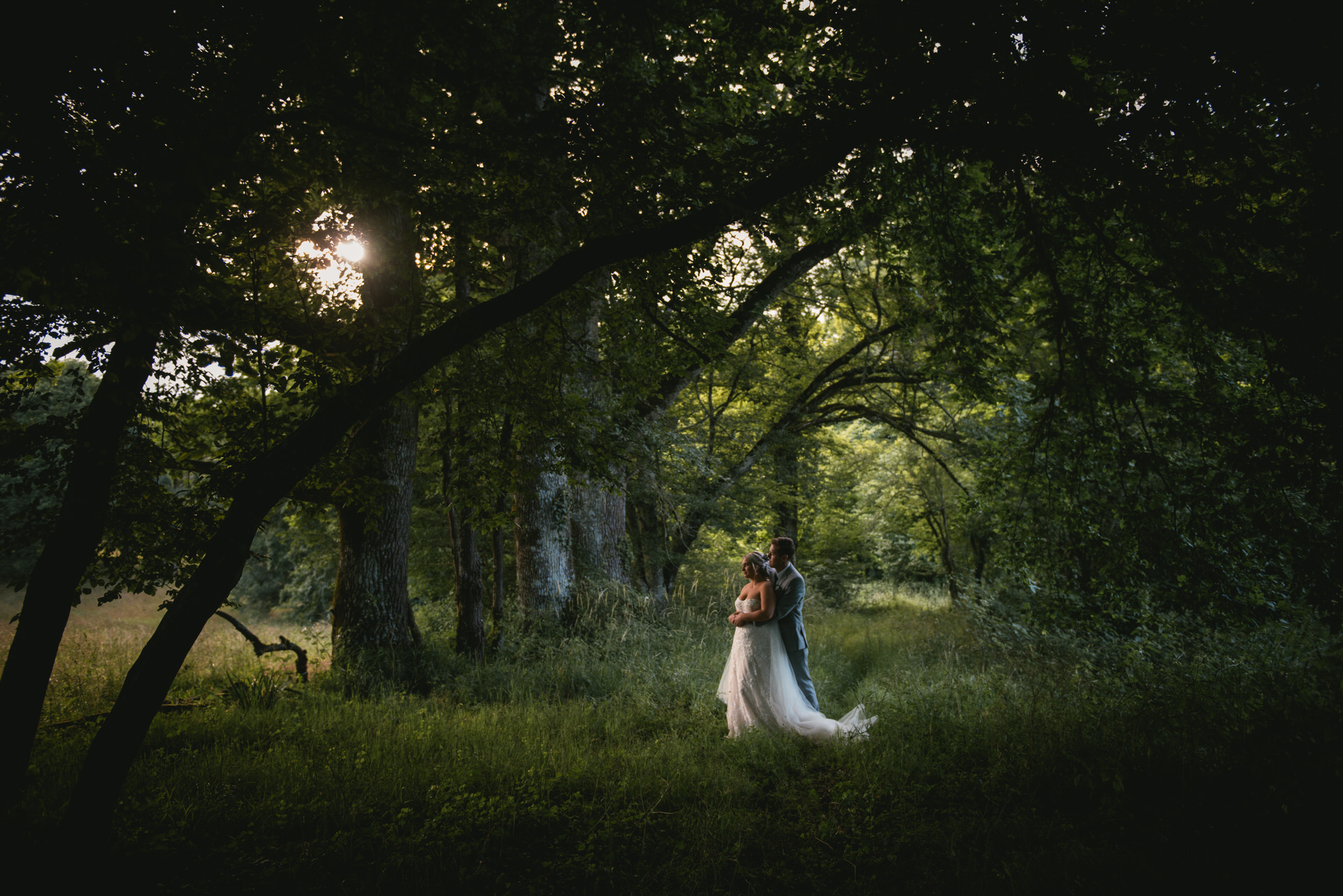 A couple stands closely embraced in a serene forest in Dordogne, France. Soft, fading light filters gently through the trees, casting a warm glow around them, creating a peaceful and intimate atmosphere.