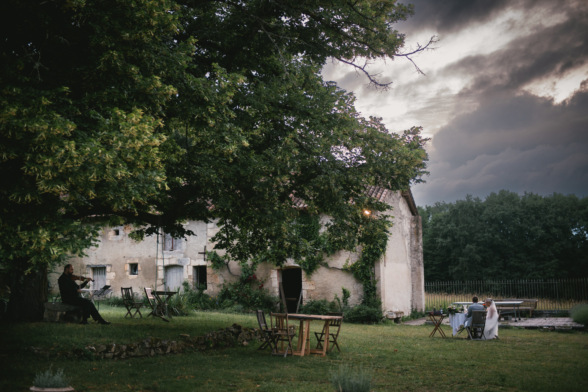 In a rustic garden in Dordogne, France, a couple shares an intimate meal at a small table under a large tree. A violinist stands nearby, playing soulful music that adds to the enchanting atmosphere.