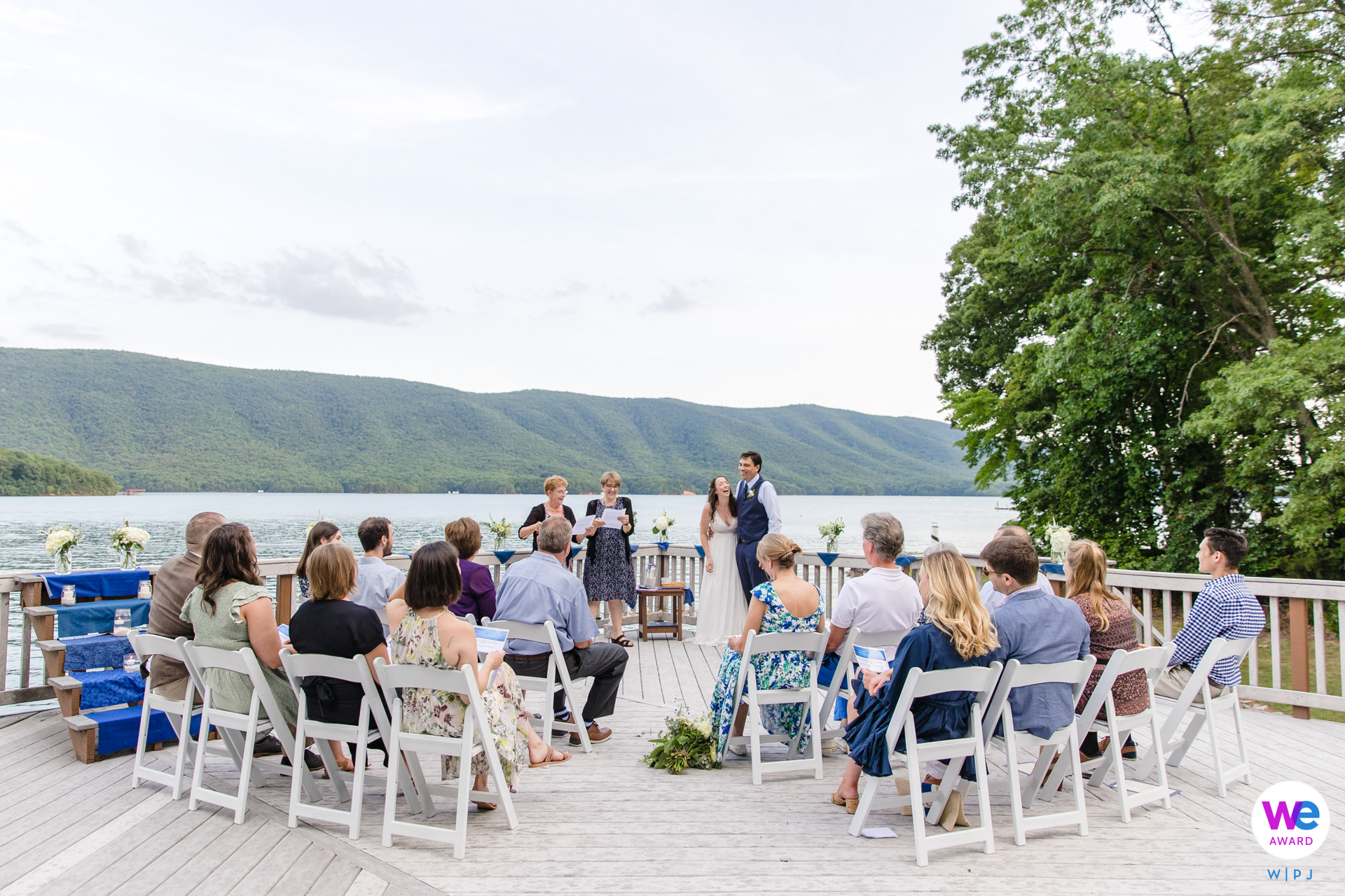 A couple exchanges vows on a deck overlooking a serene mountain lake, surrounded by loved ones.